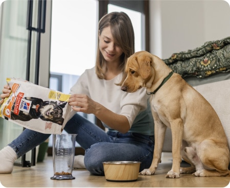 Žena sype krmivo Hill's Pet Nutrition do odměrky, zatímco ji pozorně sleduje pes.