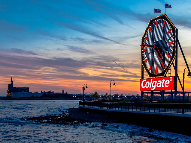 mořské pobřeží, logo Colgate v pozadí pod velkými hodinami s červenými ručičkami