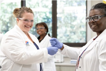 Dva vědci v laboratorních pláštích a rukavicích se usmívají a podávají si ruce v laboratorním prostředí.